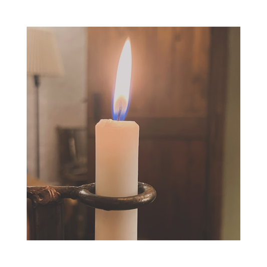 Lit white pillar candle with a steady flame in a dark metal holder, indoors with a softly blurred background