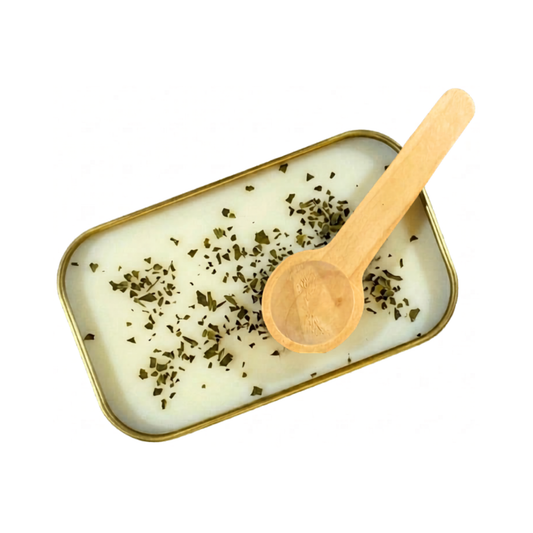 A close up view of the inside of a gold tin containing white scoopable candle wax. The surface of the wax is decorated with small dried pieces of green mint leaves. A wooden scoop rests on the edge of the tin.