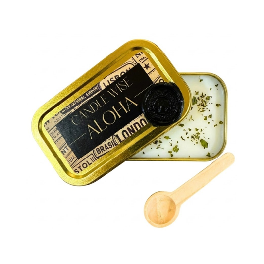 An angled photograph showing an open gold tin with the lid resting partially over the top. The label on the lid displays the name ALOHA. The white wax inside is decorated with small dried mint pieces and a wooden scoop lies nearby on a white background.
