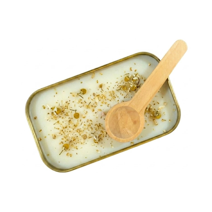 A close up view of the inside of a gold tin containing white scoopable candle wax. The surface of the wax is scattered with fine dried camomile flowers and petals. A wooden scoop rests on the edge of the tin.