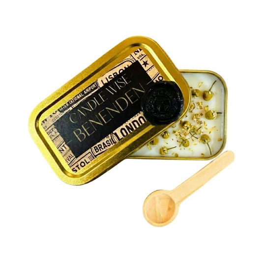 An angled photograph showing an open gold tin with the lid resting partially over the top. The label on the lid displays the name BENENDEN. The white wax inside is decorated with fine dried camomile and a wooden scoop lies nearby on a white background.