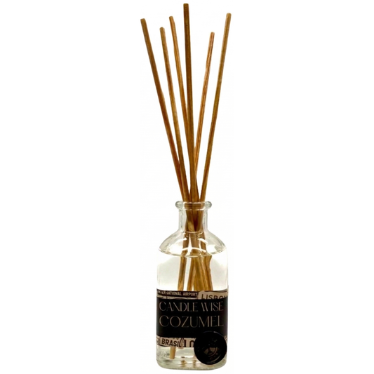 A clear glass reed diffuser bottle with a black label featuring the brand Candle Wise and the scent name COZUMEL in a serif font. Several light brown wooden reeds emerge from the narrow neck of the bottle.