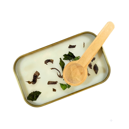 An open gold tin of Candle Wise Iguazu scoopable wax melt displaying white wax sprinkled with green jasmine tea leaves next to a small wooden spoon