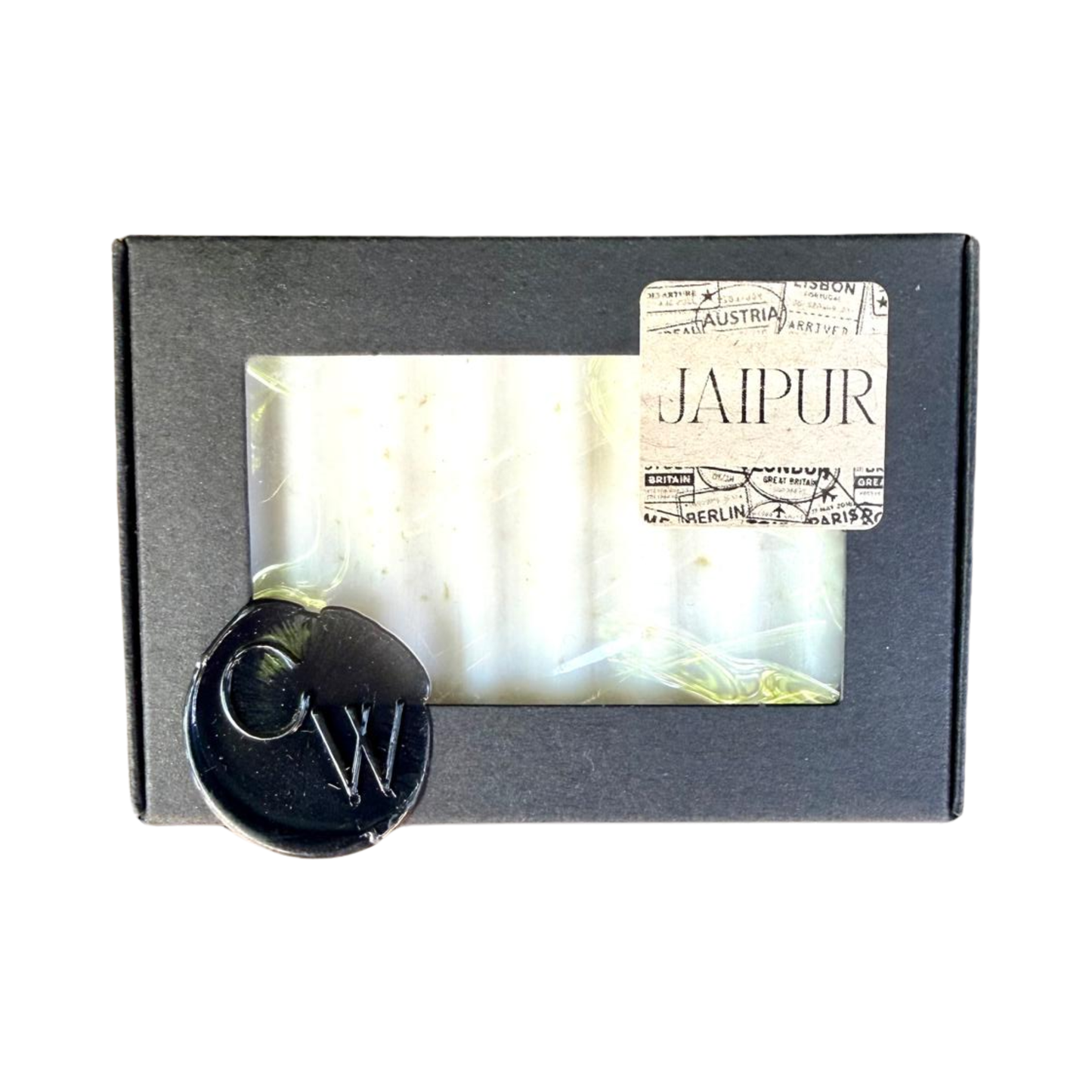 A rectangular bar of textured white soap inside a matte black cardboard box with a clear viewing window. The top right features a vintage passport style sticker reading JAIPUR, and the bottom left corner is detailed with a black CW wax seal.