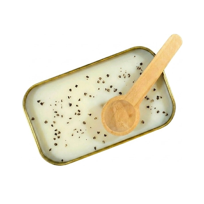 A close up view of the inside of a gold tin containing white scoopable candle wax. The surface of the wax is lightly sprinkled with fine black tea speckles. A wooden scoop rests on the edge of the tin.