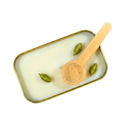 A close up view of the inside of a gold tin containing white scoopable candle wax. The surface of the wax is decorated with three green cardamom pods spread out. A wooden scoop rests on the edge of the tin.