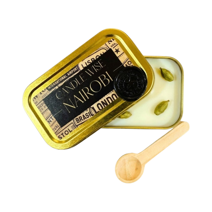 An angled photograph showing an open gold tin with the lid resting partially over the top. The label on the lid displays the name NAIROBI. The white wax inside features cardamom pods and a wooden scoop lies nearby on a white background.