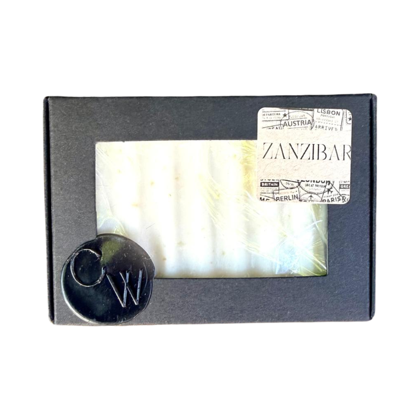 A rectangular bar of textured white soap packaged in a matte black cardboard box with a rectangular window. A vintage style travel stamp sticker with the word ZANZIBAR is on the top right corner, and a black wax seal with the letters CW is on the bottom left corner.